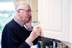 Forgetful Senior Man With Dementia Looking In Cupboard At Home