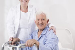 A senior sits down with his hands on a walker as a woman stands behind him with her hand on his shoulder, assisting with managing ALS symptoms.