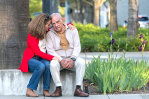 An adult child of a senior experiencing behavioral changes after a brain injury wraps her arm around him as they sit outside.