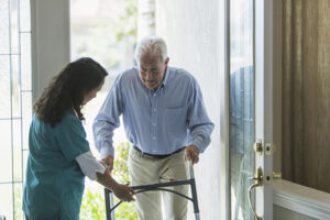 A senior man, helped by his caregiver, uses a walker to get into his home after returning from a doctor’s appointment to learn if he has Parkinson’s or dementia with Lewy bodies.